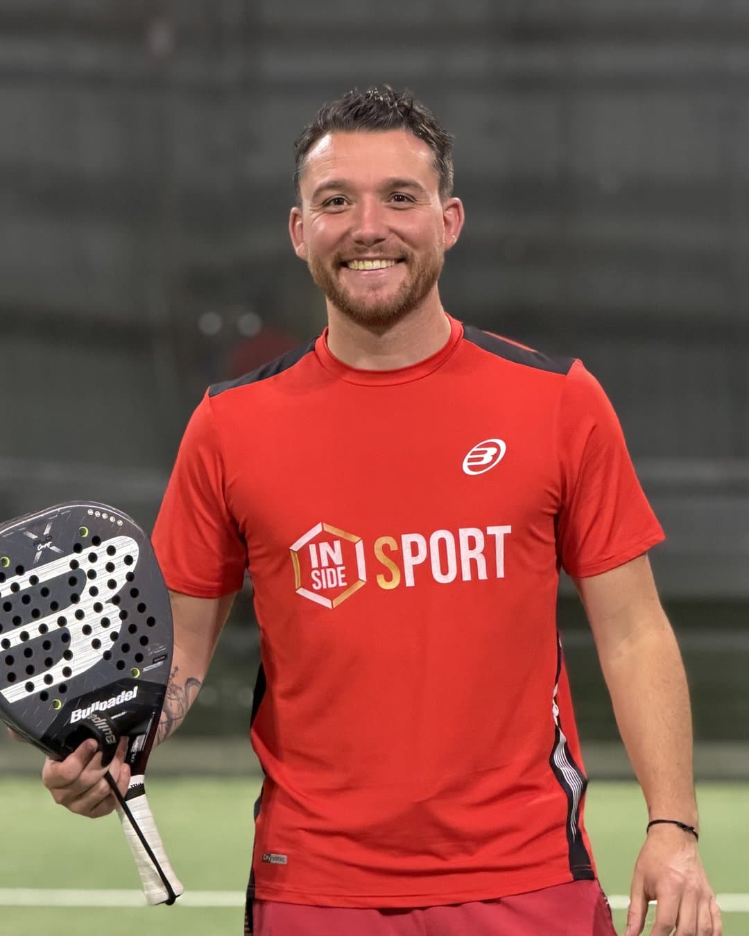 Inside Sport padel coach, racket in hand, smiling at the net of a court in Reims