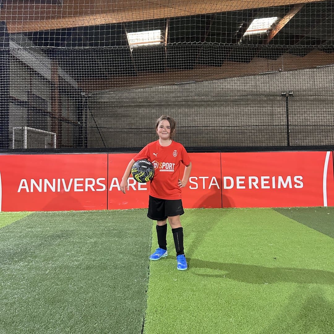 Child in an Inside Sport shirt, ball under arm, in front of the Stade de Reims Birthday banner